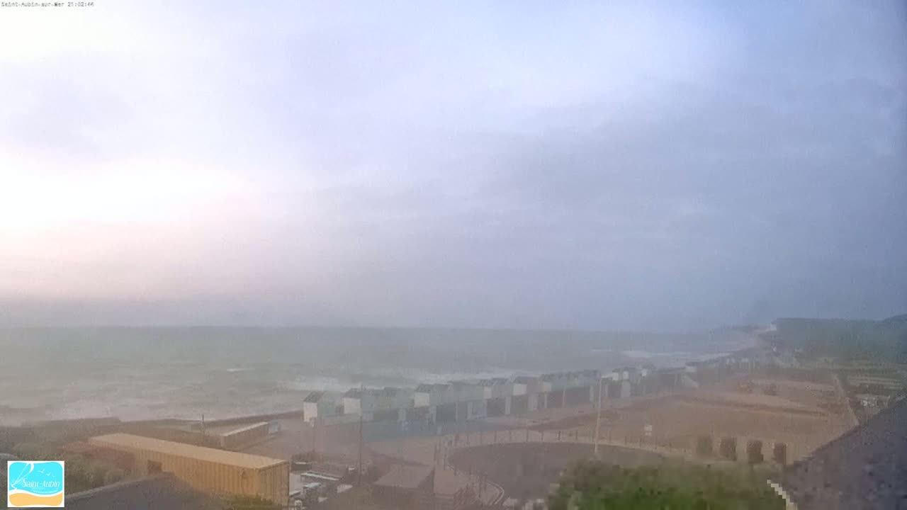 A hazy, overcast day shows a choppy sea with waves crashing near a beachline with several low buildings.