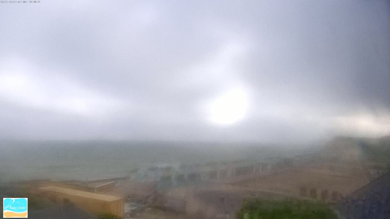 A hazy sun shines through an overcast sky over a blurry coastal landscape with buildings and a body of water.