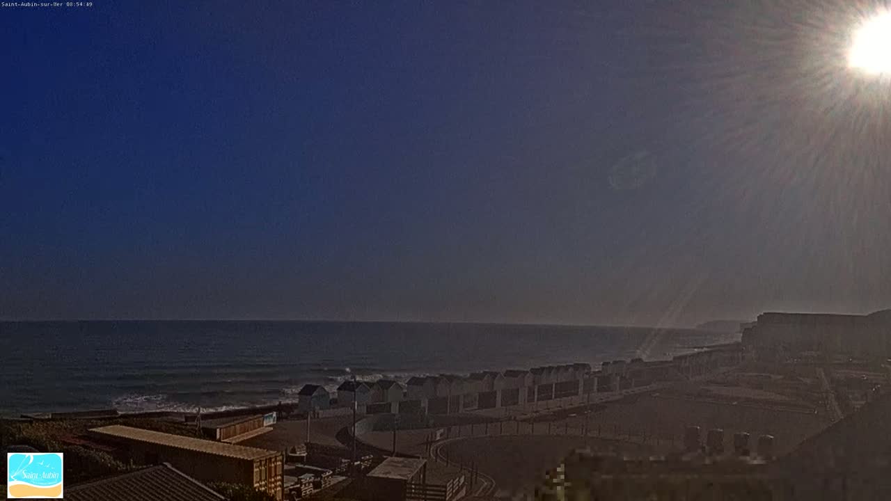 A sunny day shows a row of beach huts next to a calm ocean, with a cliff visible in the distance.
