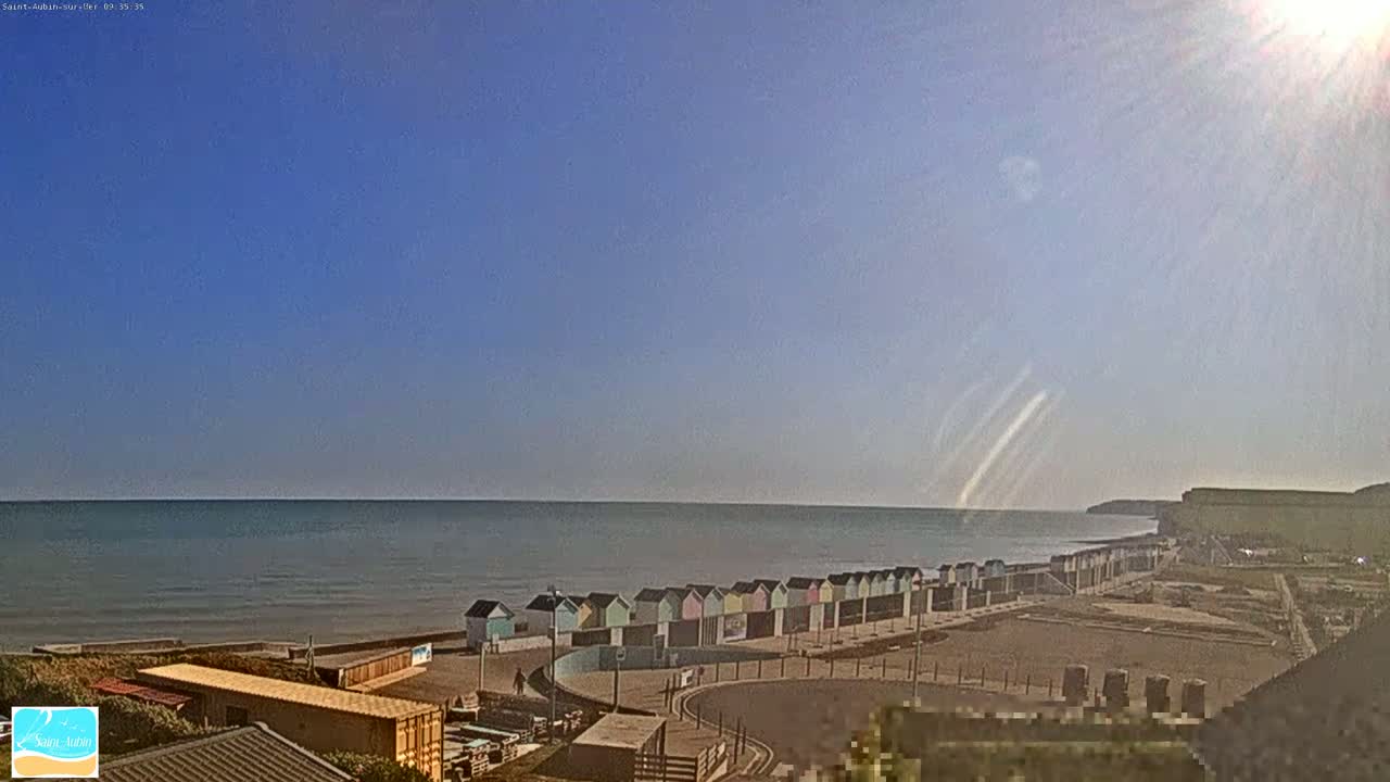 A sunny day at the beach shows a row of colorful beach huts next to a calm ocean with a distant cliff in the background.