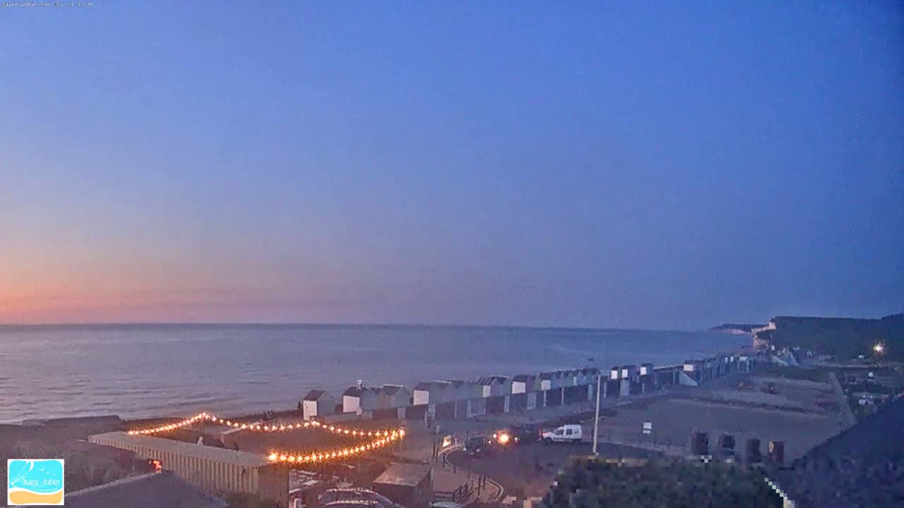 A row of beach huts is illuminated by string lights at dusk, overlooking a calm sea and distant cliffs under a clear twilight sky.