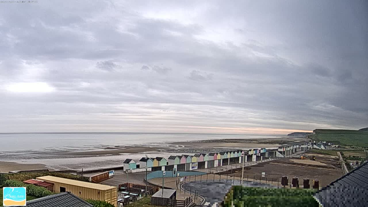 A row of colorful beach huts lines a sandy shore under a cloudy sky, with a calm sea extending to a distant cliff.