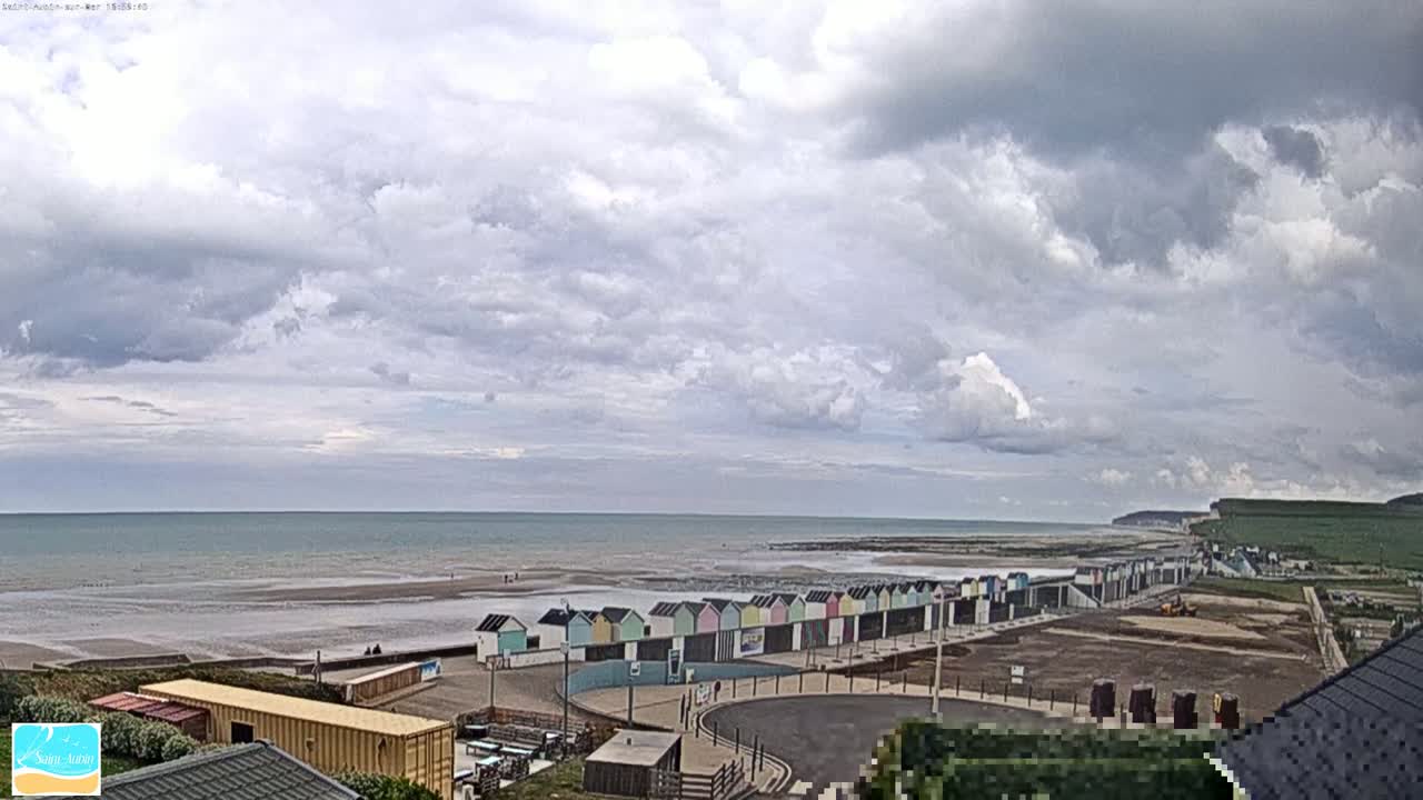 A row of colorful beach huts lines a sandy beach, under a partly cloudy sky, with a calm ocean extending to the horizon and cliffs visible in the distance.
