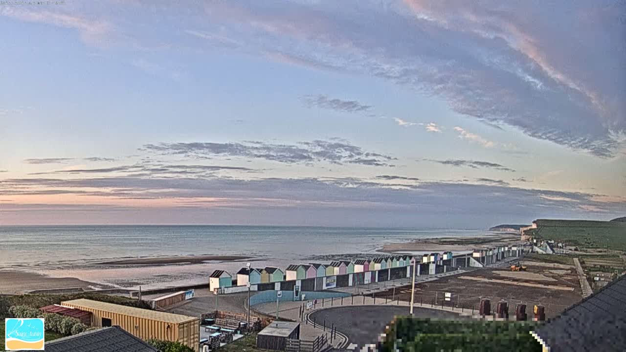 A row of colorful beach huts sits on a sandy beach overlooking a calm ocean under a partly cloudy sunset sky.