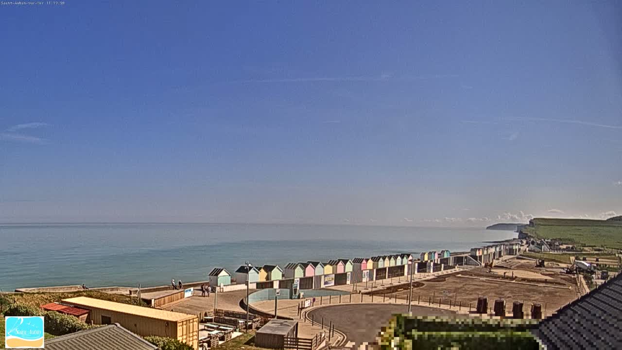 Under a clear blue sky, a calm ocean stretches to the horizon, where white cliffs are visible in the distance, beside a row of colorful beach huts situated along a paved walkway.