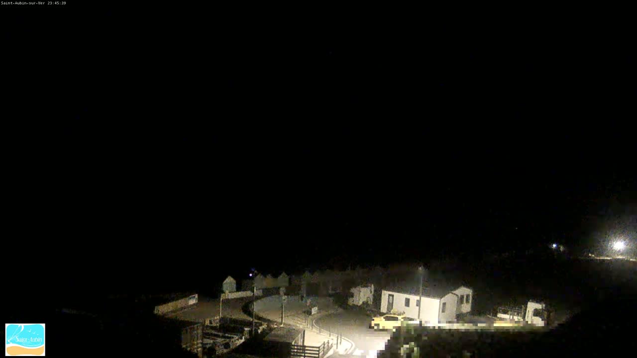 A nighttime, dimly lit coastal scene shows a few buildings and a car parked near a road curving around a small area.