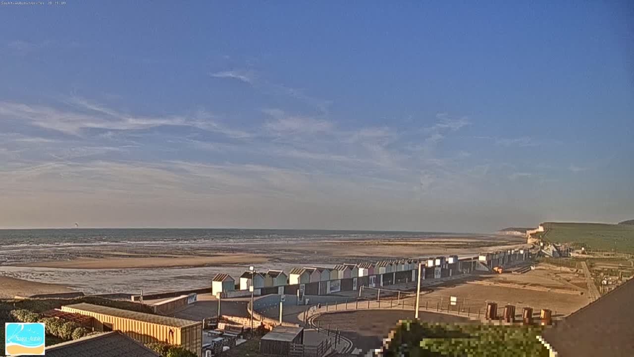 A row of colorful beach huts lines a sandy beach under a mostly clear, blue sky, with a calm ocean in the background and white cliffs visible in the distance.