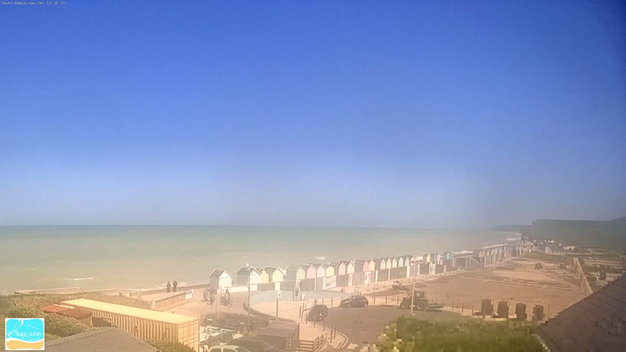 A hazy, sunny day overlooks a beach with a row of colorful beach huts, a calm sea, and a parking area.