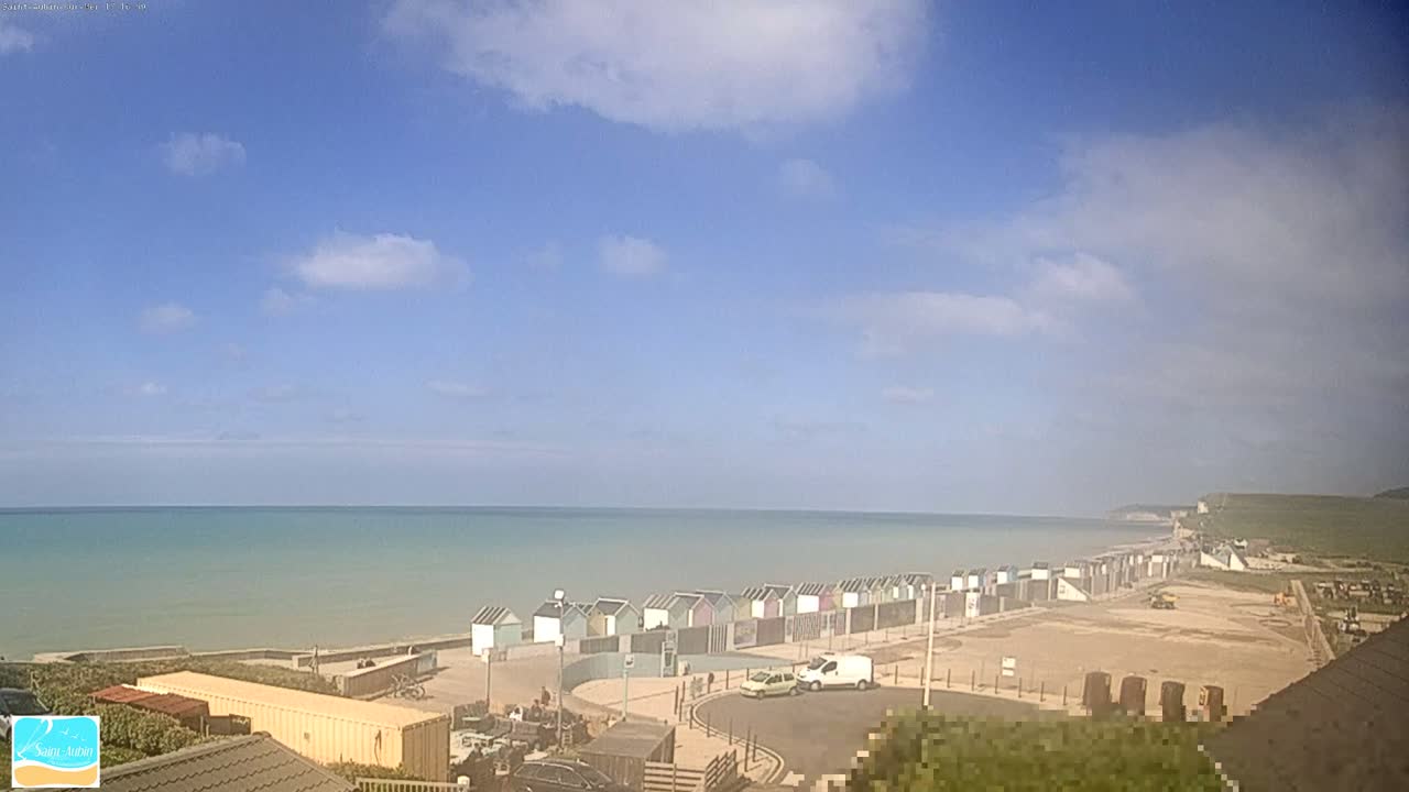 A row of beach huts sits on a sandy beach overlooking a calm, light-blue sea under a mostly clear, bright sky with some scattered clouds.