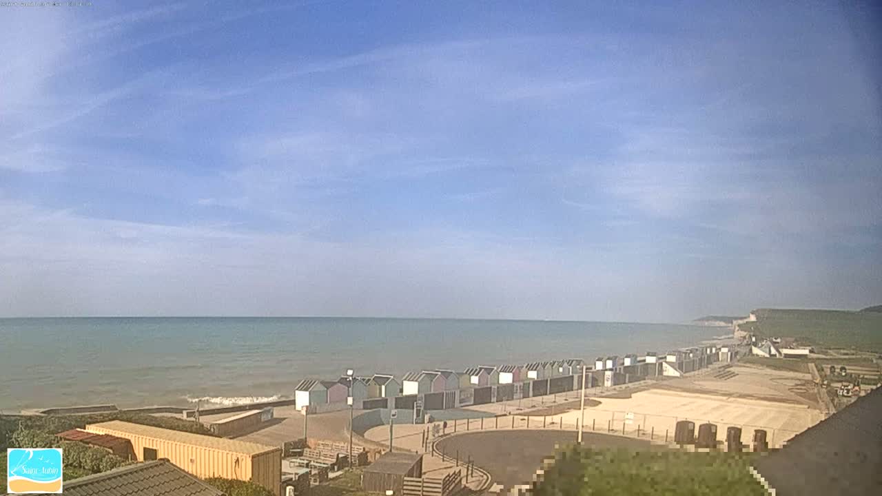 A row of colorful beach huts lines a sandy shore next to a calm, blue ocean under a mostly clear sky.