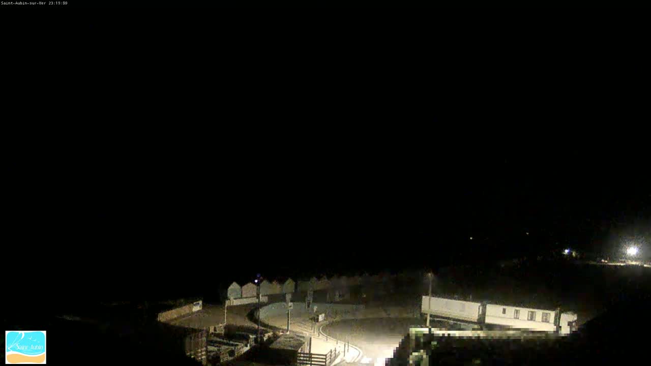 A nighttime, dimly lit view of a coastal area shows a curved road leading to a row of beach huts under a dark sky.