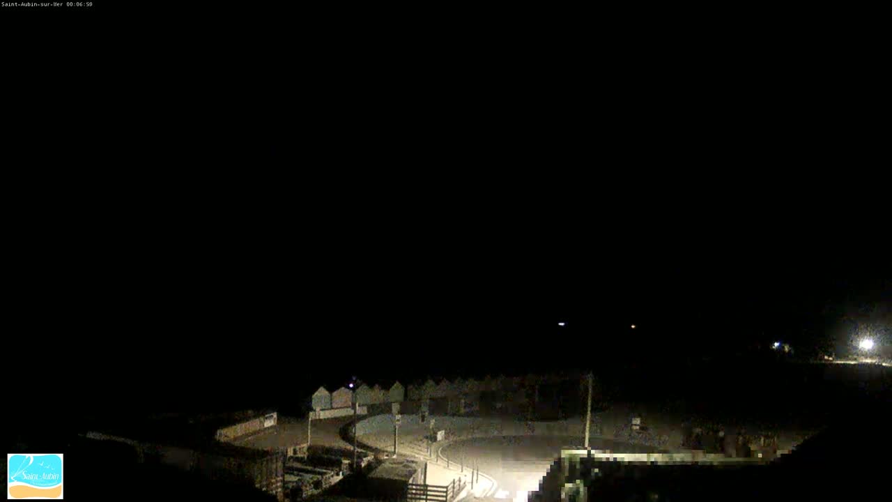 A nighttime, high-angle view shows a dark, mostly empty beach area with a few dimly lit buildings and a curved roadway.