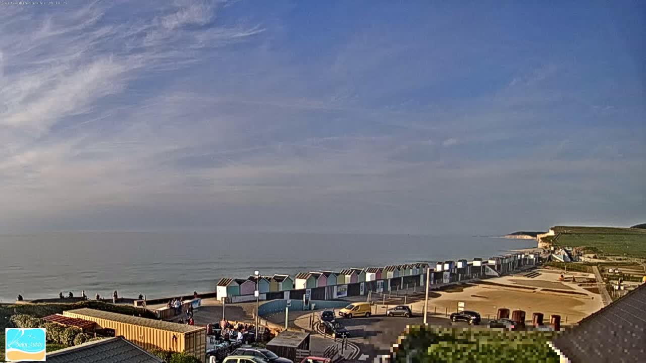 A beach with colorful beach huts, a calm ocean, and a partly cloudy sky is viewed from an elevated position.