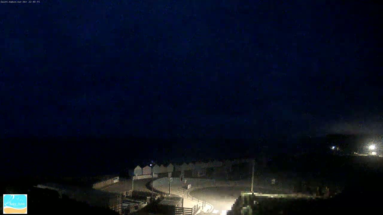 A nighttime, dimly lit coastal scene shows a curved road leading to a row of beach huts near the dark ocean under a clear, dark sky.