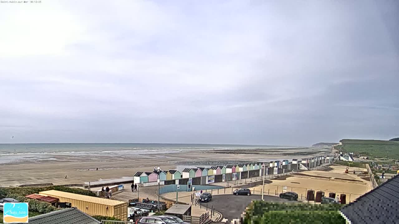A wide sandy beach stretches to a calm ocean under a mostly cloudy sky, with a row of colorful beach huts and a parking area visible closer to the viewer.
