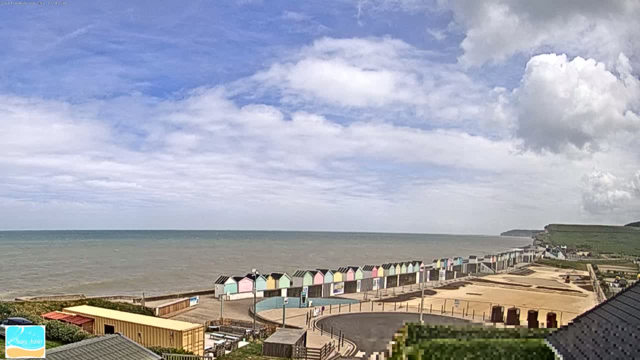 A row of colorful beach huts sits along a calm, gray ocean under a partly cloudy sky, with a sandy area and cliffs visible in the background.