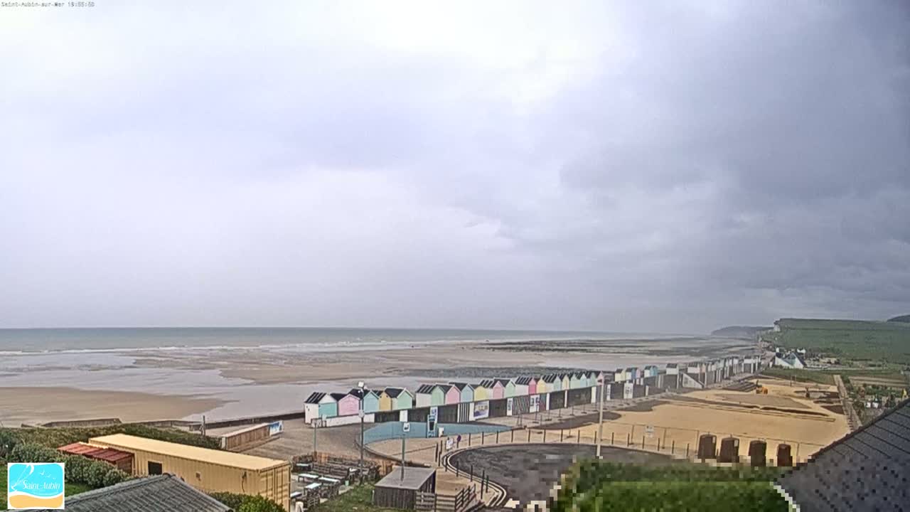 A row of colorful beach huts sits on a sandy beach next to a calm, gray ocean under an overcast sky.