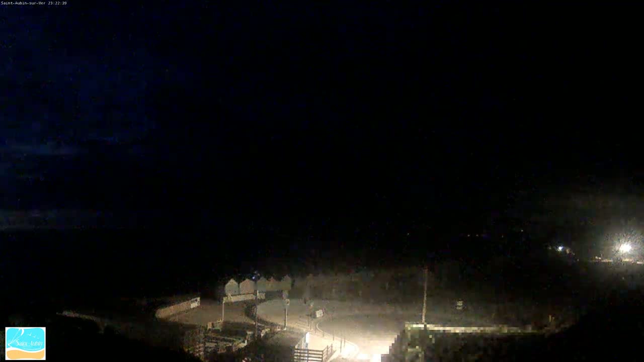A nighttime, dark view of a coastal area with dimly lit buildings and a roadway near the water under a mostly clear sky.