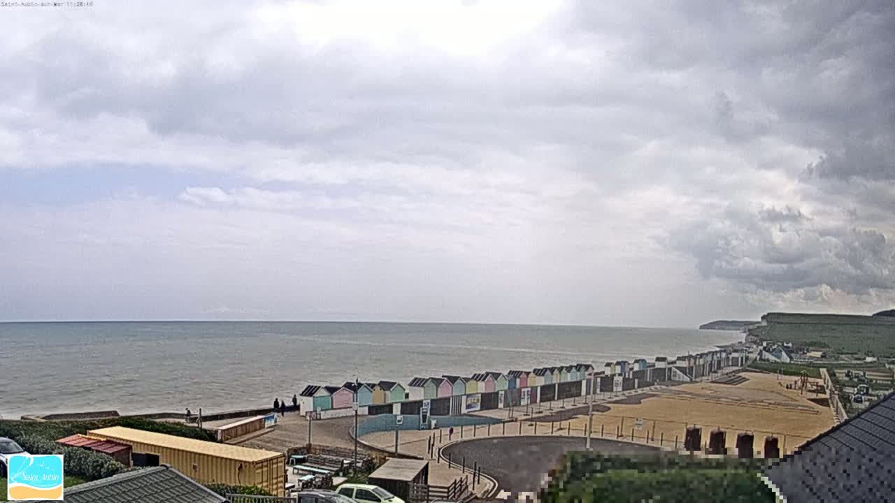 A row of colorful beach huts sits on a sandy beach overlooking a calm sea under a cloudy sky.