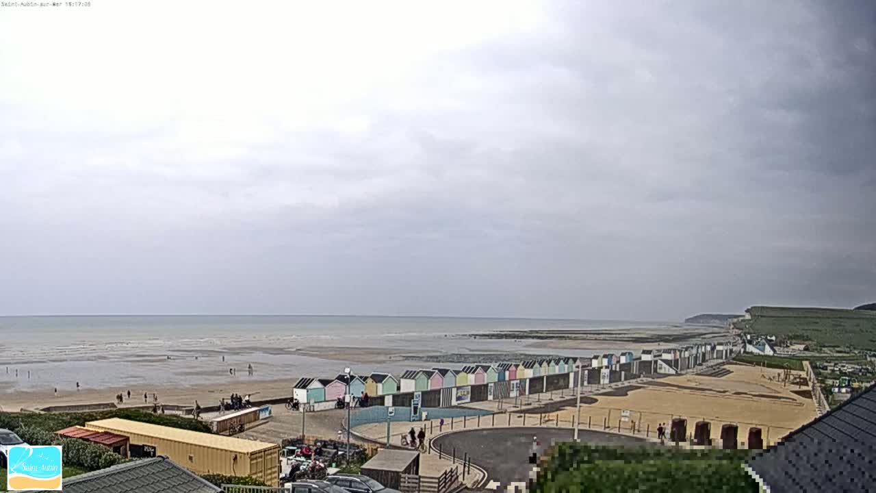 A wide shot of a beach with colorful beach huts under an overcast sky shows a calm ocean and sparsely populated sandy shore.