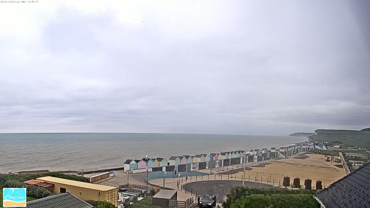 A row of colorful beach huts sits on a sandy beach next to a calm, gray sea under an overcast sky.