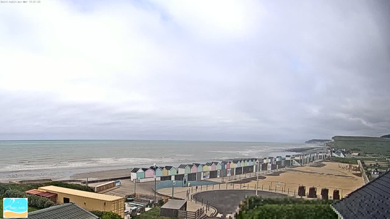 A row of colorful beach huts sits on a sandy beach overlooking a calm, gray sea under an overcast sky.