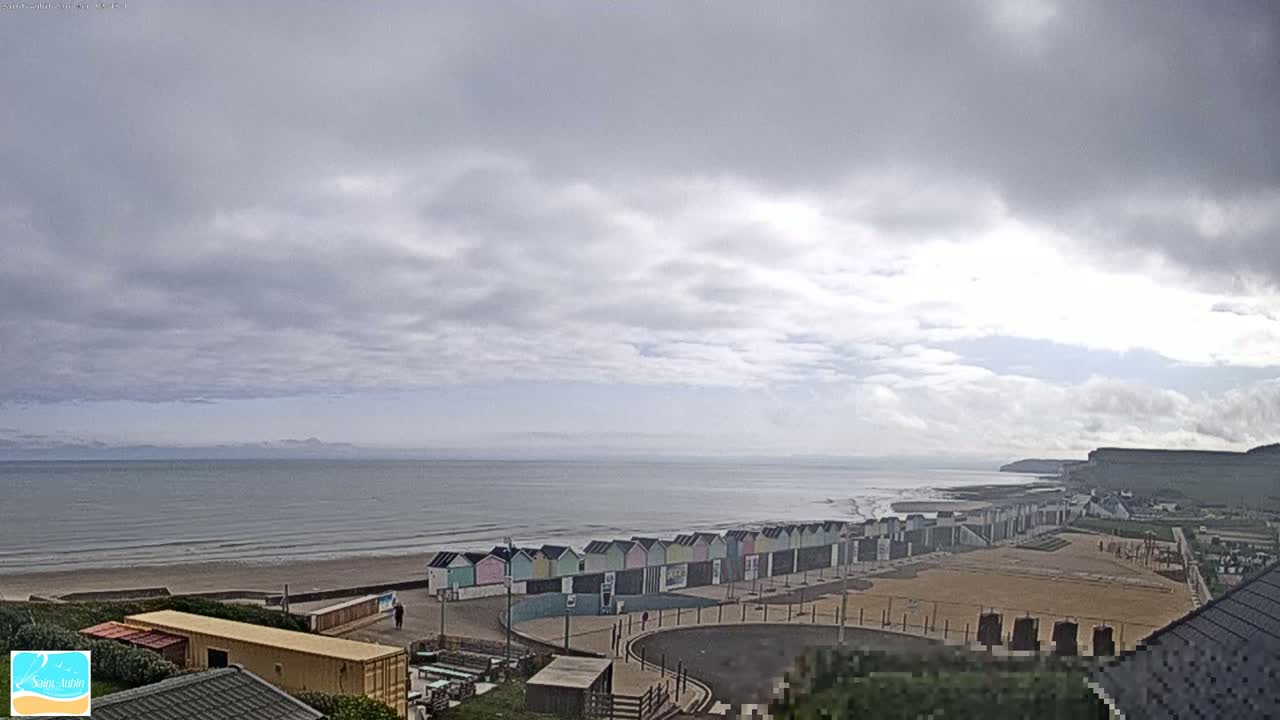 A mostly cloudy day shows a long row of colorful beach huts next to a wide sandy beach and calm ocean, with cliffs visible in the distance.