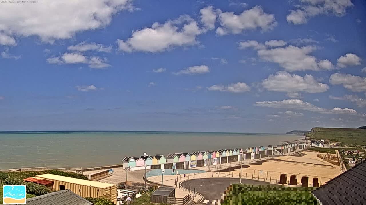 A row of colorful beach huts sits on a sandy beach overlooking a calm, gray-green sea under a partly cloudy blue sky.