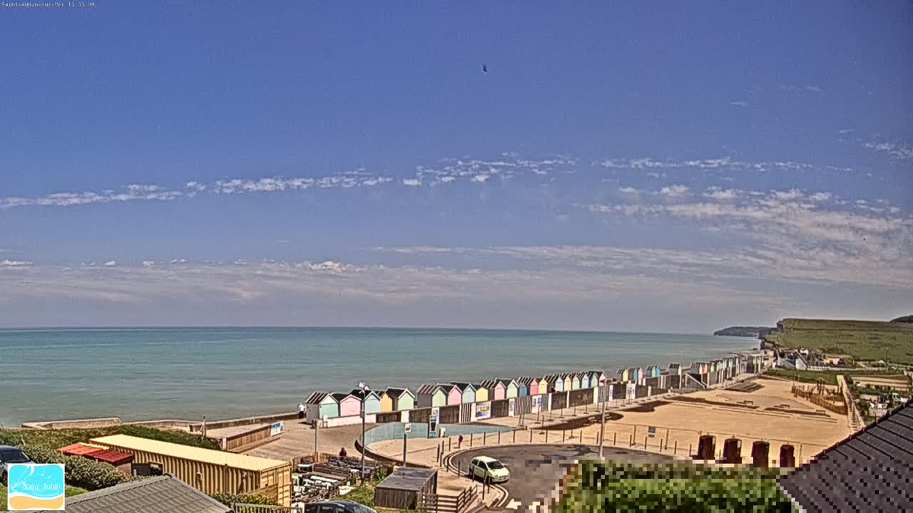 A row of colorful beach huts sits on a sandy beach overlooking a calm, light-blue ocean under a mostly sunny sky with scattered clouds.