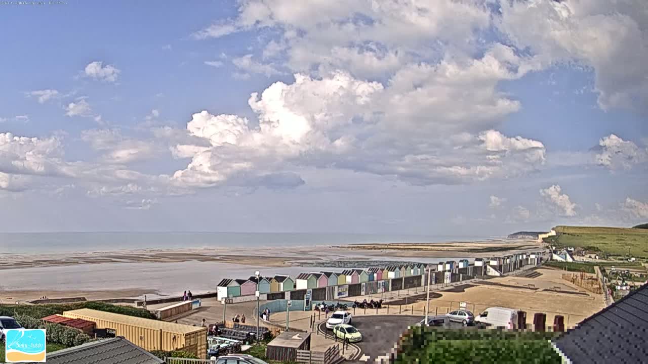 A partly cloudy sky overlooks a beach with colorful beach huts, a parking lot, and a distant cliff, under a mostly sunny sky.