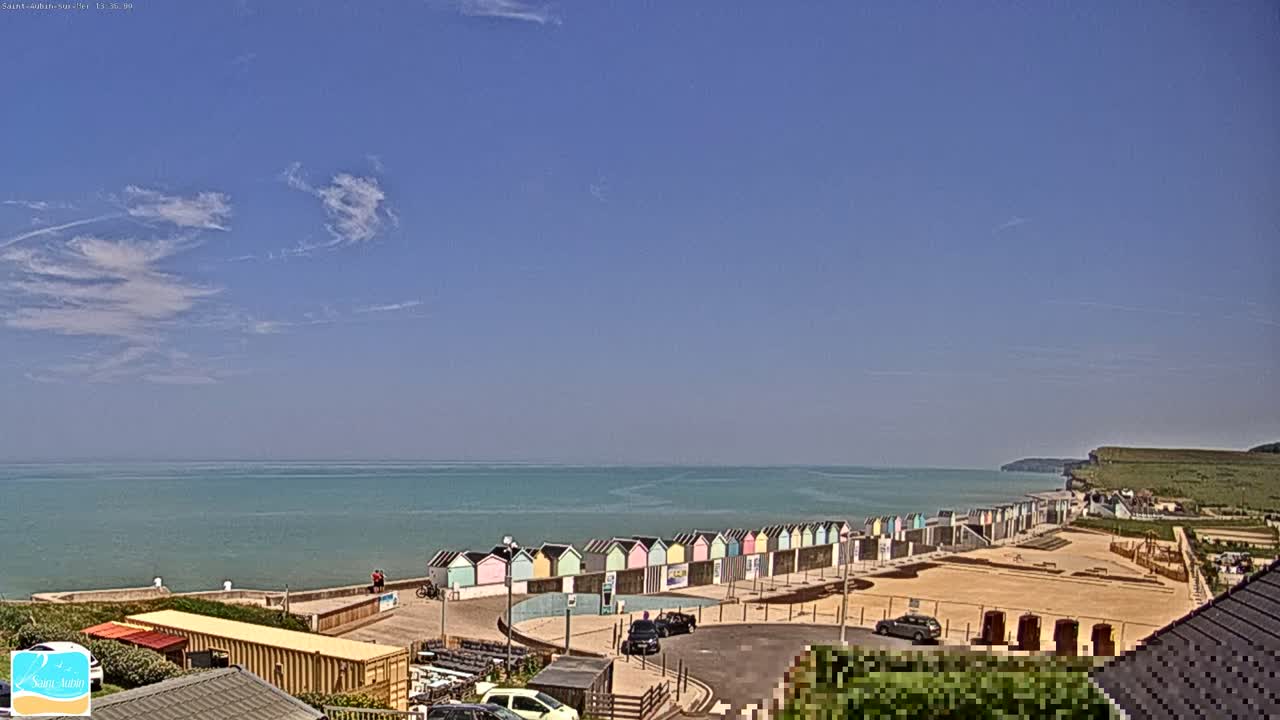 A row of colorful beach huts sits on a sandy beach overlooking a calm ocean under a mostly clear blue sky.
