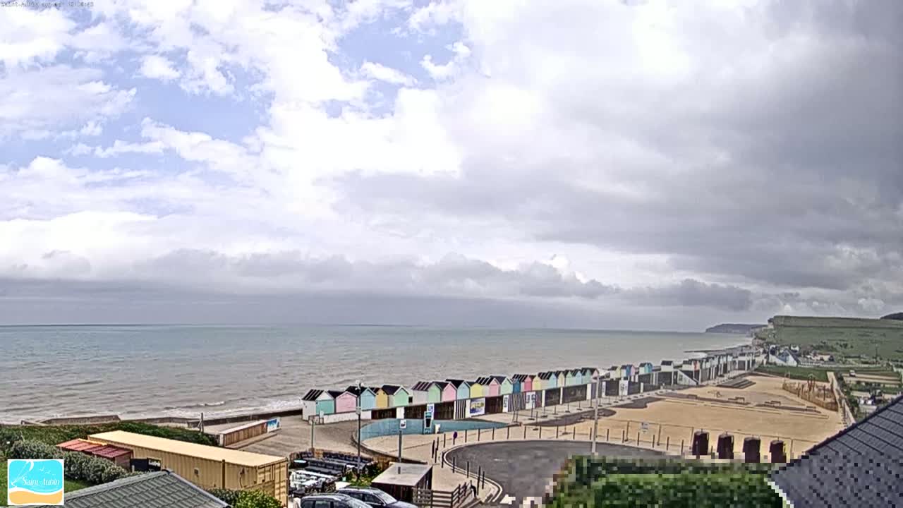 A row of colorful beach huts lines a sandy beach next to a calm, gray-blue sea under a mostly cloudy sky.