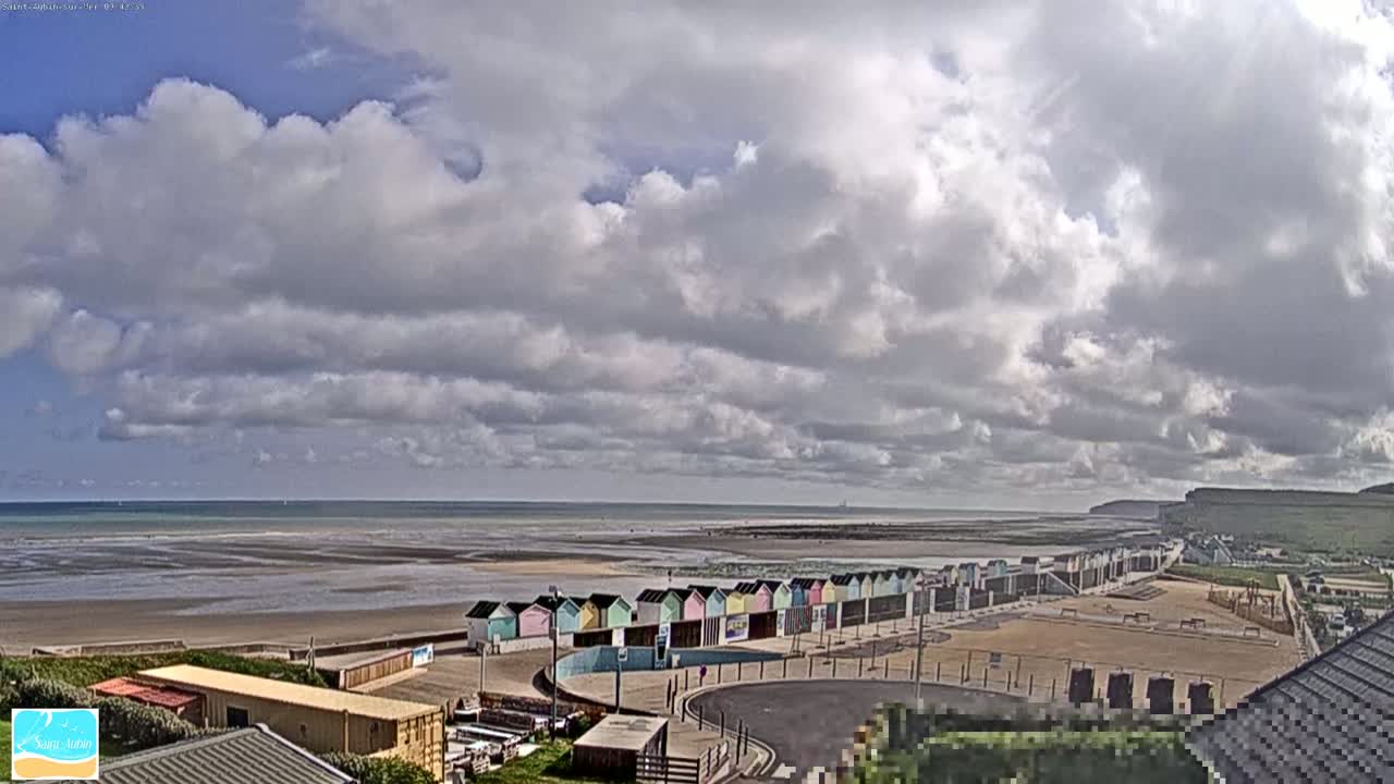A mostly cloudy sky overlooks a wide sandy beach with a row of colorful beach huts, a calm ocean, and a distant cliff.