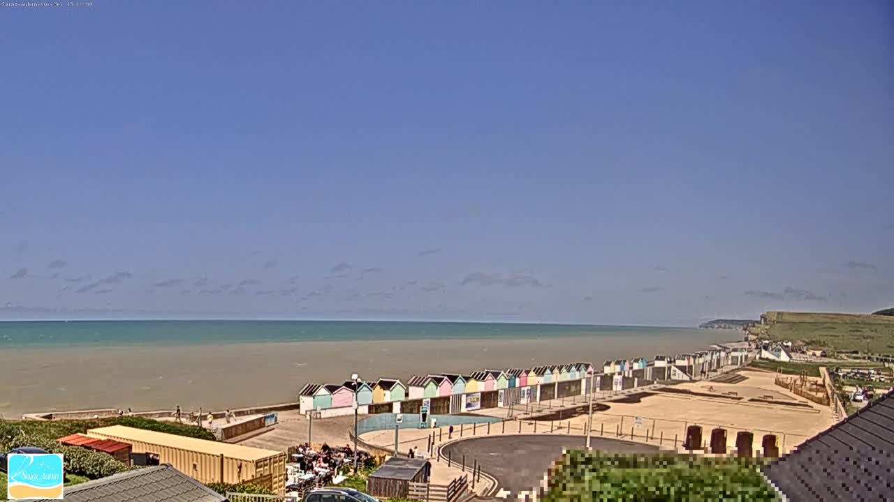 A row of colorful beach huts sits on a sandy beach overlooking a calm sea under a mostly clear sky.