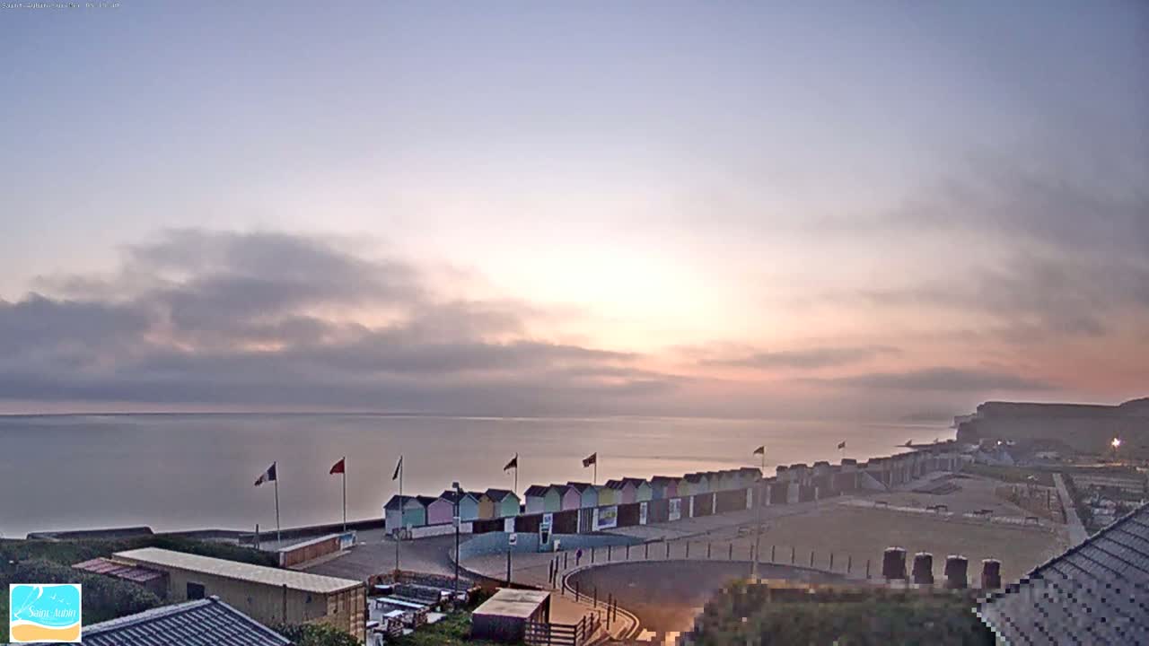 A sunrise over a calm sea with pastel-colored beach huts and a row of flags visible in the foreground.