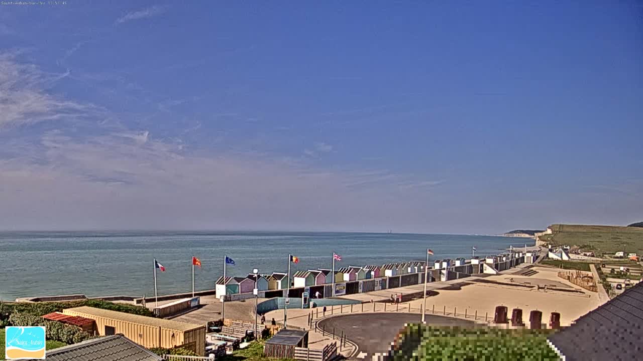 Under a mostly clear blue sky, a row of colorful beach huts sits along a sandy beach overlooking a calm ocean and distant white cliffs.