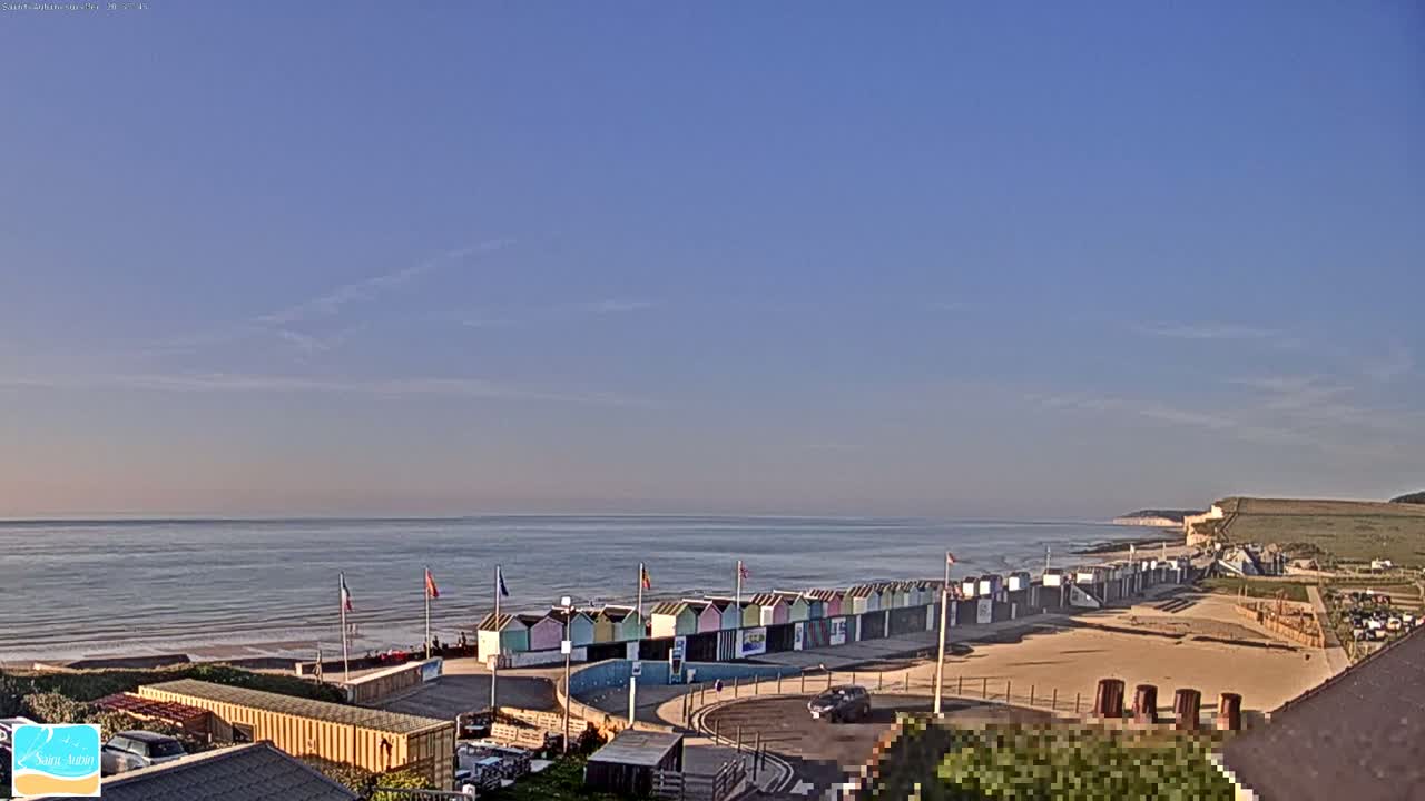 A row of colorful beach huts sits on a sandy beach overlooking a calm ocean under a clear, light blue sky.