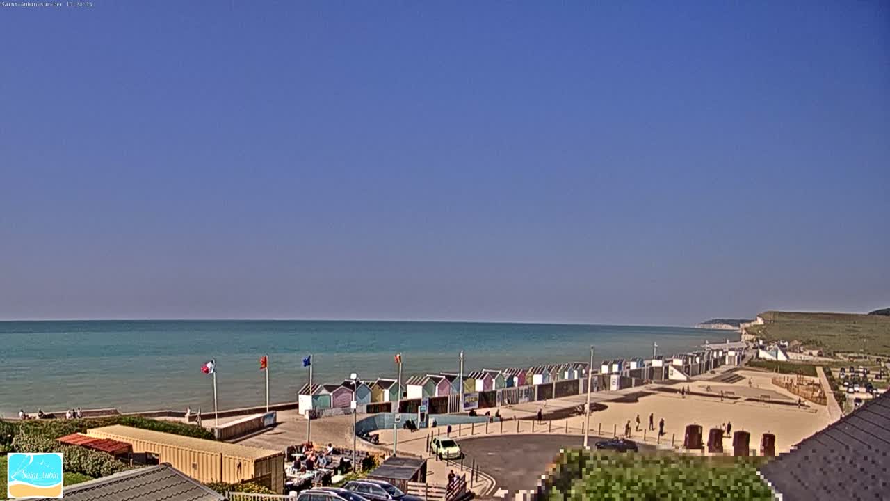 A calm, sunny day at a beach with colorful beach huts lining the shore, a few people scattered on the sand, and white cliffs visible in the distance.