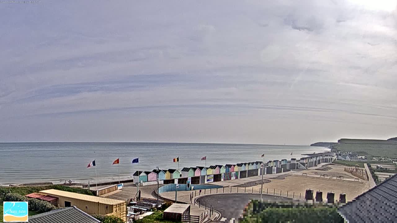 A row of colorful beach huts lines a sandy area next to a calm ocean under a mostly cloudy sky.