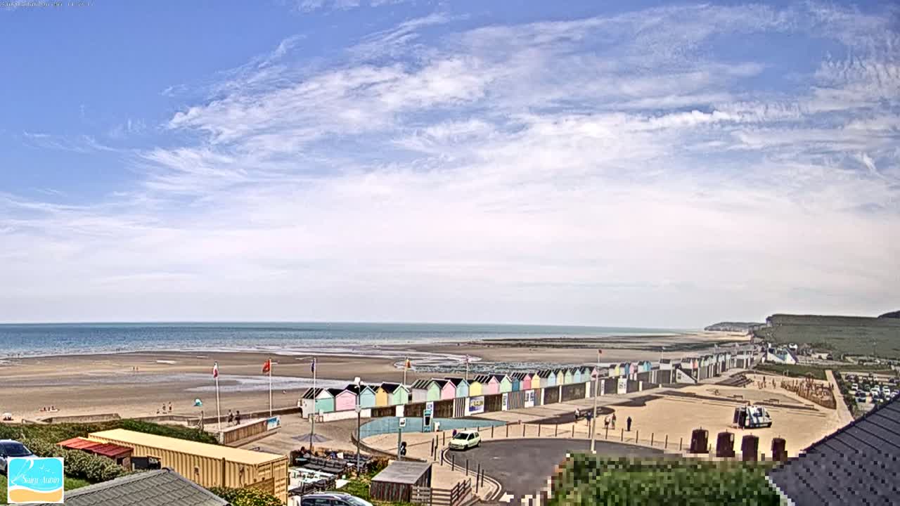 A row of colorful beach huts sits on a sandy beach next to a calm ocean under a partly cloudy sky.