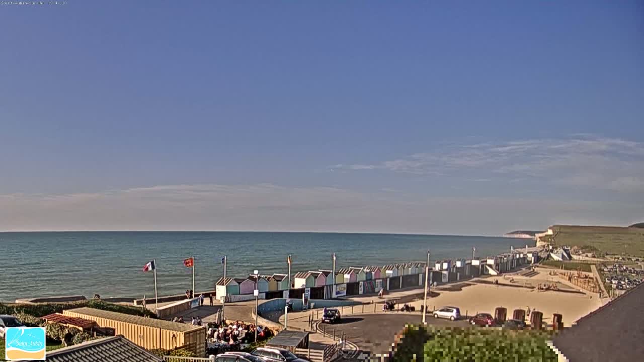 A row of colorful beach huts sits on a sandy beach next to a calm ocean under a mostly clear blue sky.