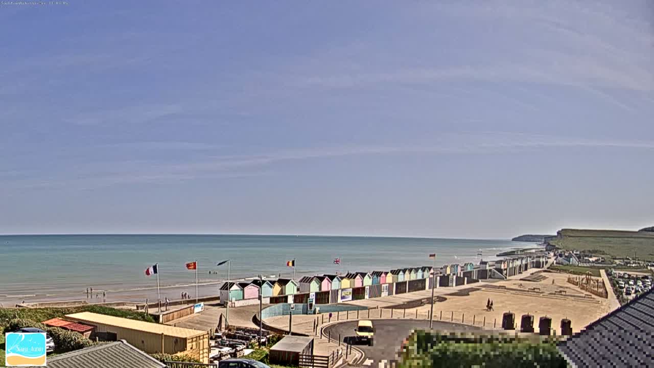 A row of colorful beach huts sits on a sandy beach overlooking a calm ocean under a mostly clear sky.