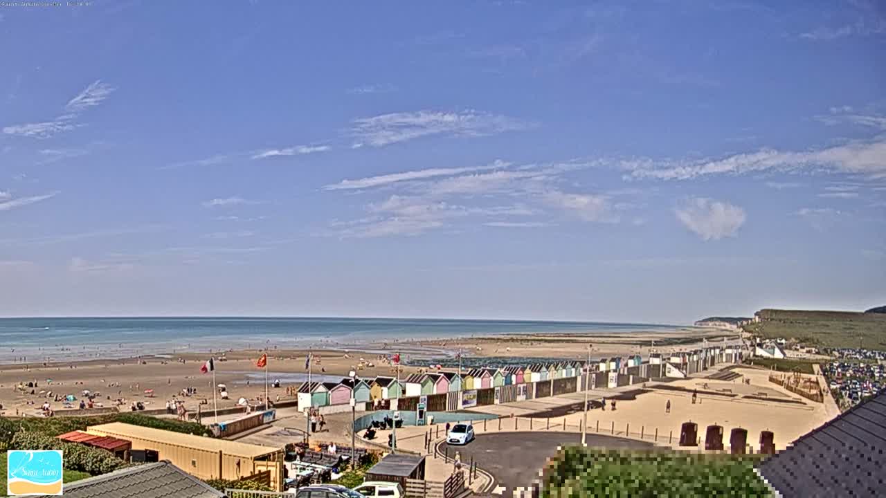 A sandy beach with colorful beach huts stretches along a calm ocean under a mostly clear, sunny sky.