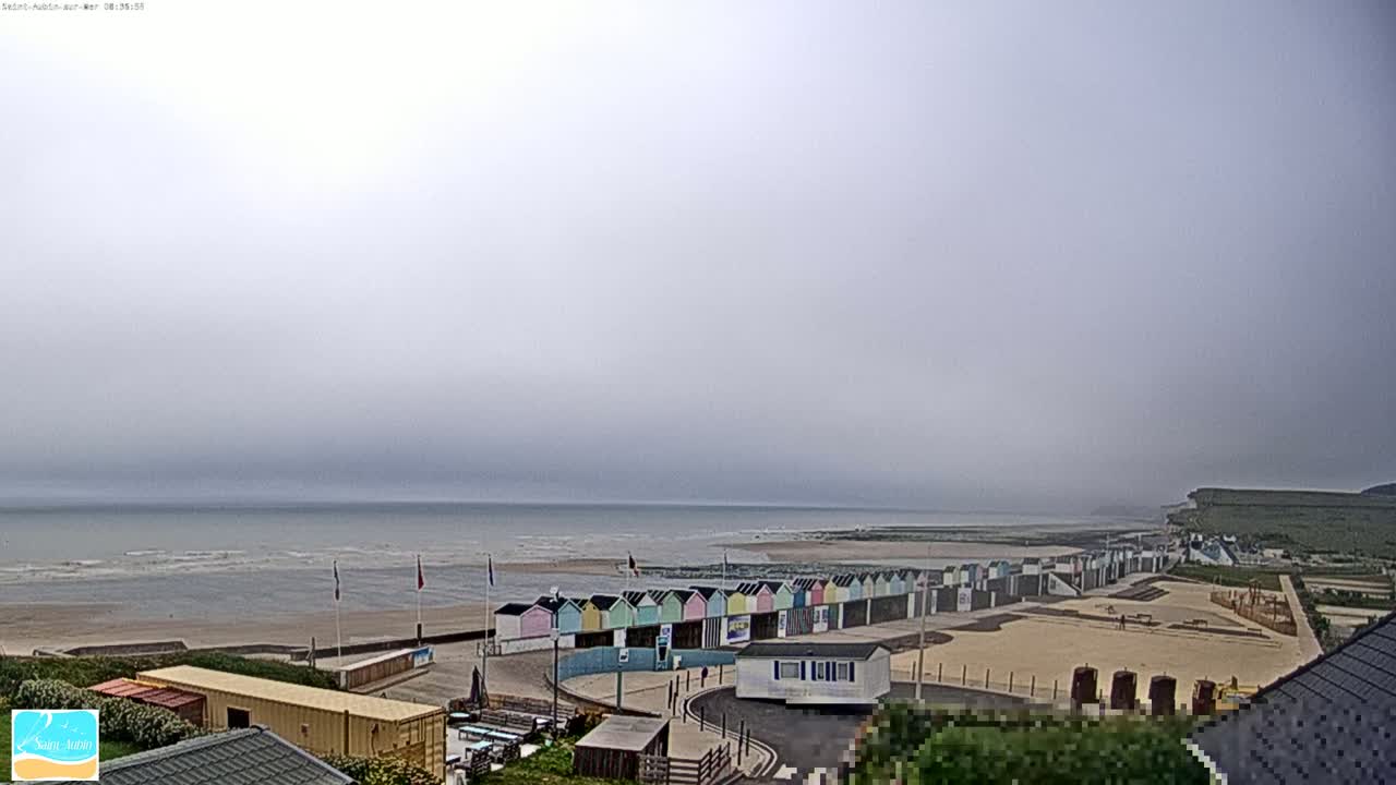 A row of colorful beach huts lines a sandy beach beside a calm, grey sea under an overcast sky.