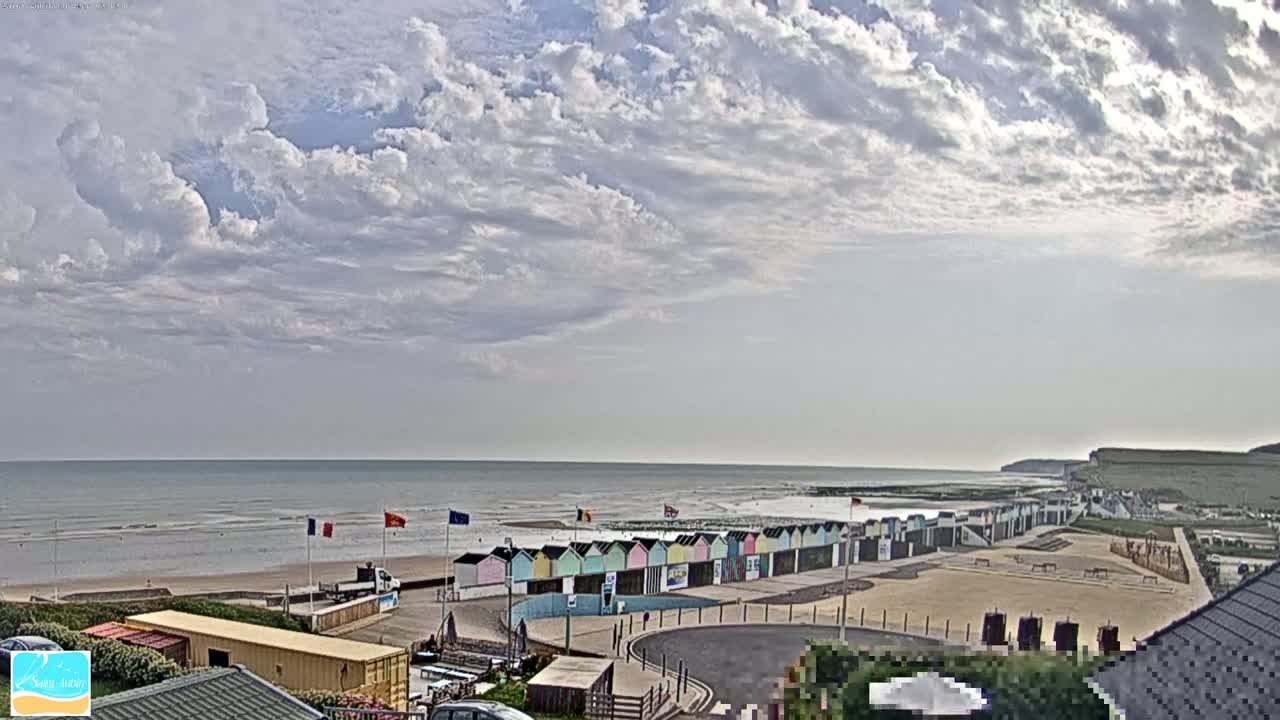 A row of colorful beach huts sits on a sandy beach overlooking a calm sea under a partly cloudy sky.