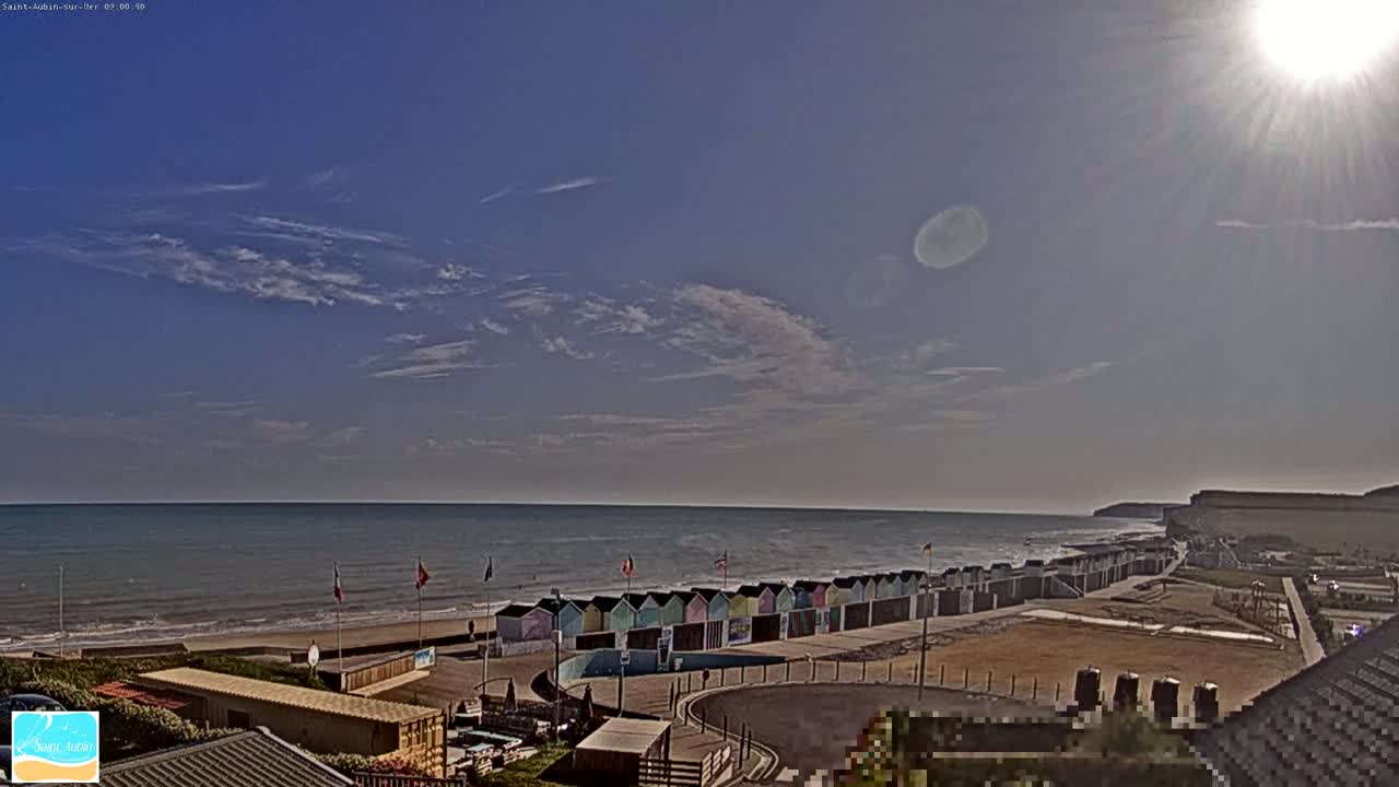 A sunny day with a mostly clear sky shows a beach with colorful beach huts and a calm ocean, viewed from a high vantage point.