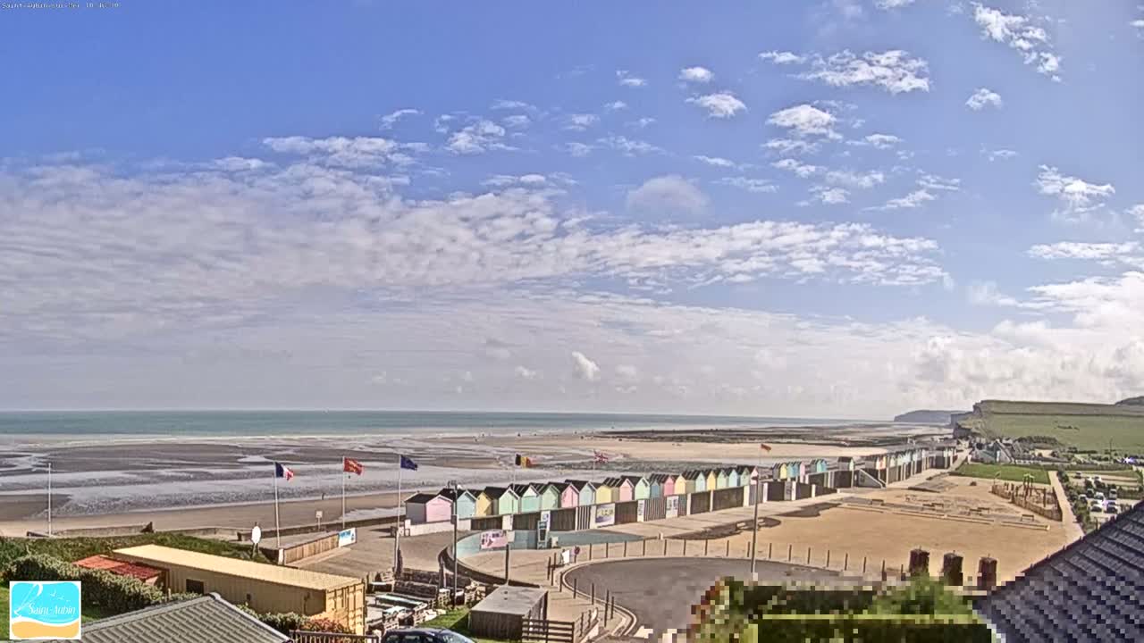 A mostly sunny day shows a beach with pastel-colored beach huts, a wide sandy area, and a calm ocean under a partly cloudy sky.