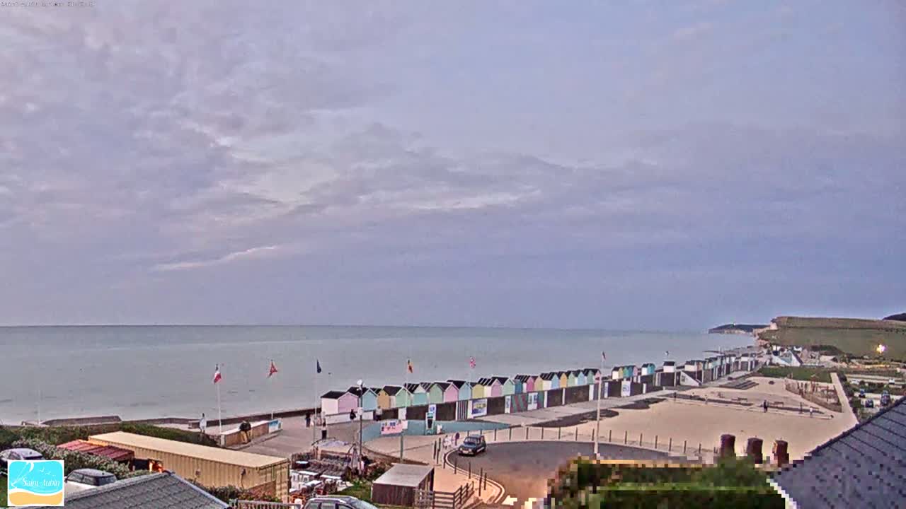 A mostly cloudy day reveals a beach with colorful beach huts, a calm sea, and white cliffs in the distance.