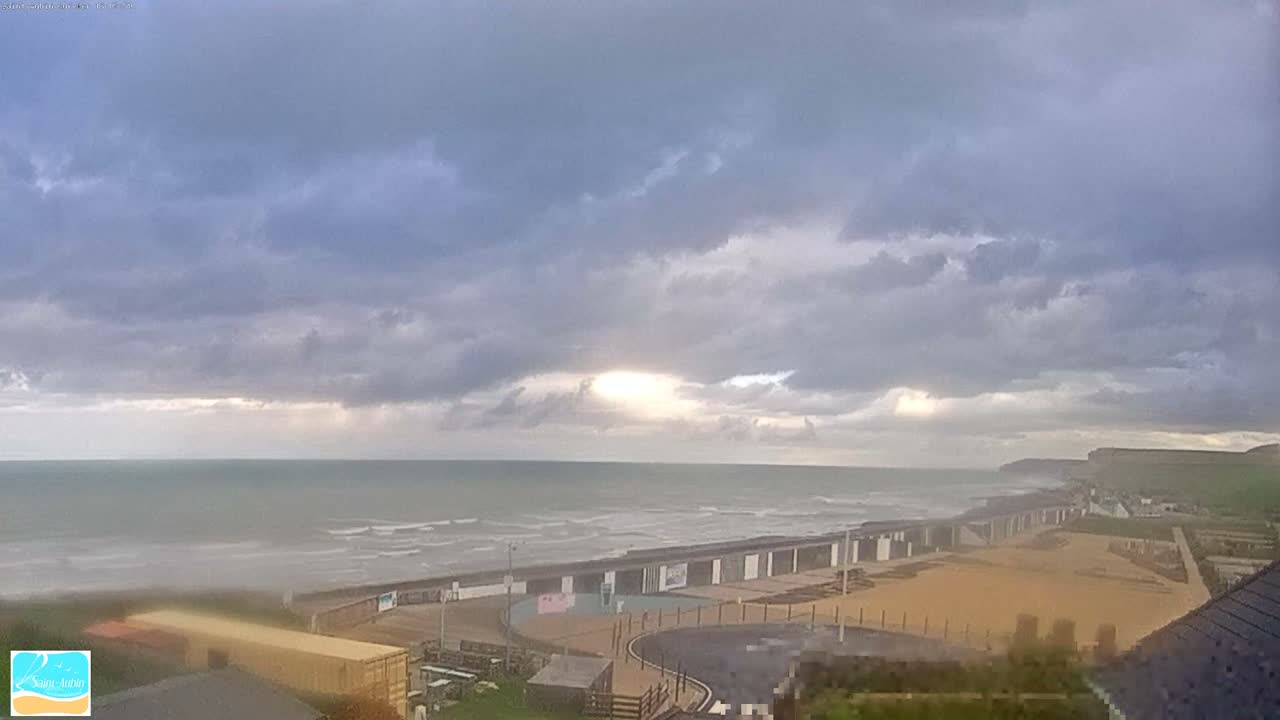 A mostly cloudy day reveals a beach with colorful beach huts, a calm sea, and white cliffs in the distance.