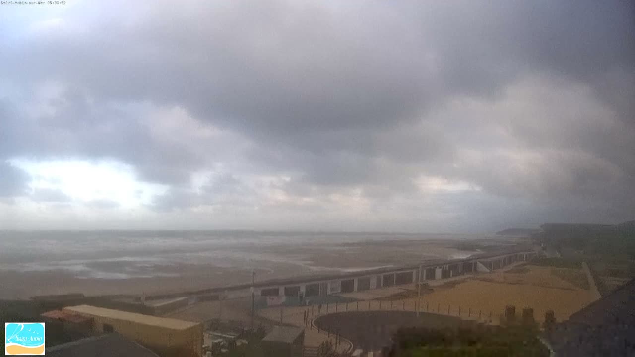 A wide coastal view shows a choppy sea washing onto a sandy beach alongside a long promenade and buildings, all beneath a heavily overcast and grey sky.